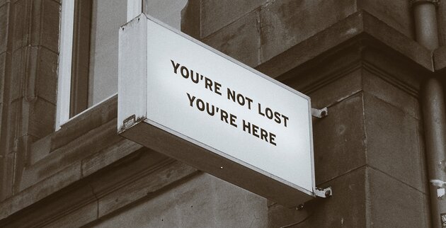 Picture of a sign placed on the corner of a building saying "You're not lost, you're here.". Picture in sepia by Eileen Pan.